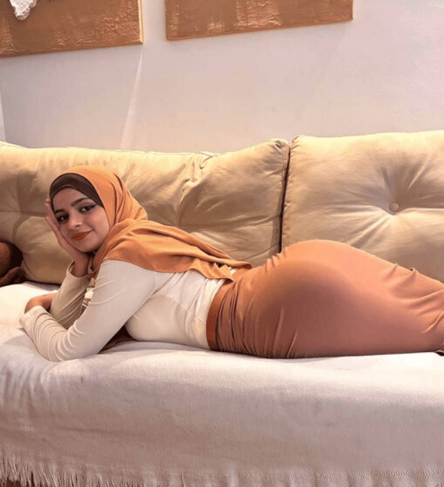 Woman wearing a peach hijab and beige outfit lying on a beige sofa, propped on her elbows and smiling at the camera.