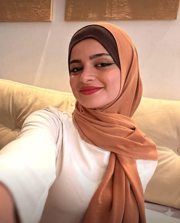 Woman wearing a peach hijab and white top taking a selfie on a beige sofa, smiling at the camera.