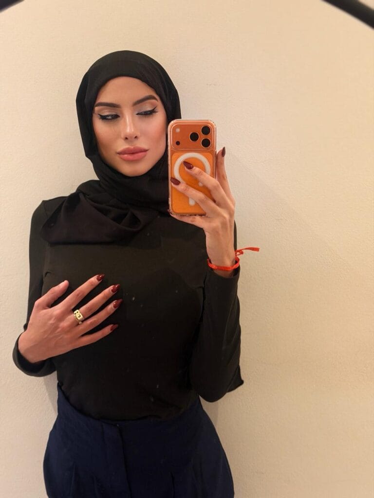 Woman in a black hijab and top taking a mirror selfie, beige wall behind.