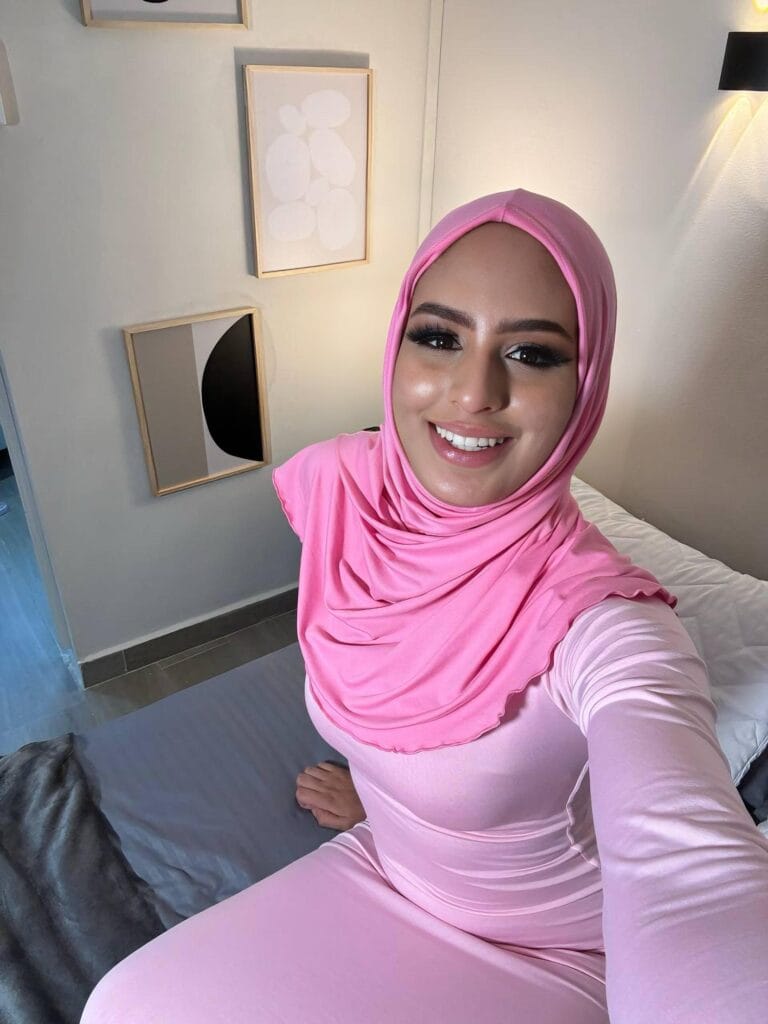 Smiling woman in a pink hijab and matching outfit taking a selfie in a softly lit room with abstract wall art behind her.