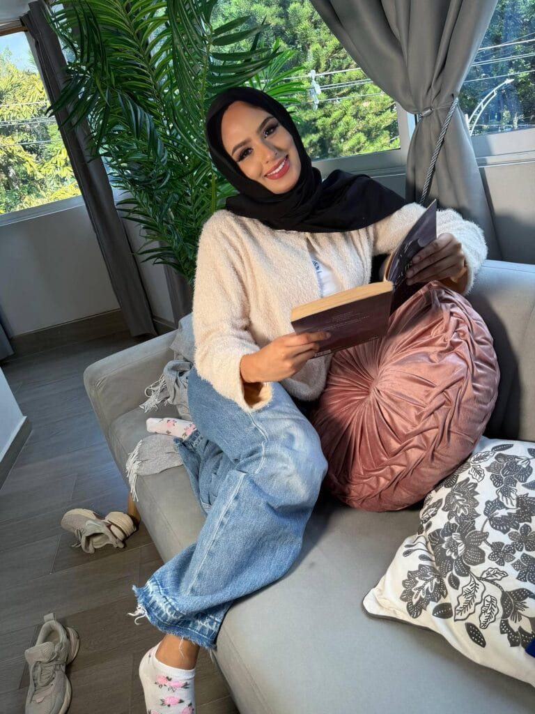 Smiling woman in a black hijab and beige cardigan sits on a gray sofa reading a book, with leafy plants in the background.