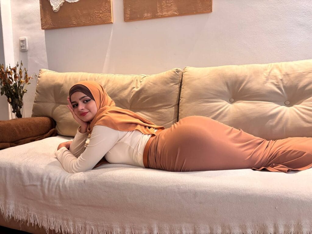 Woman in a tan hijab and beige outfit lying on a cream sofa, propped on her elbow and smiling at the camera.