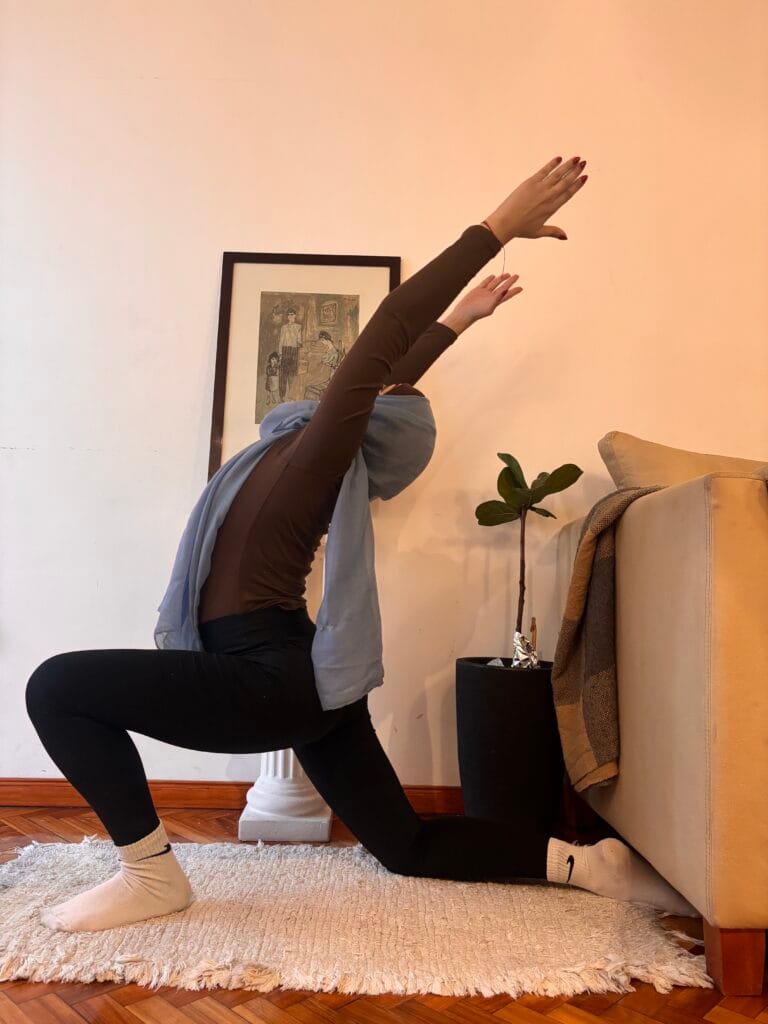 Woman in a hijab practicing yoga on a rug in a living room, arms raised overhead.