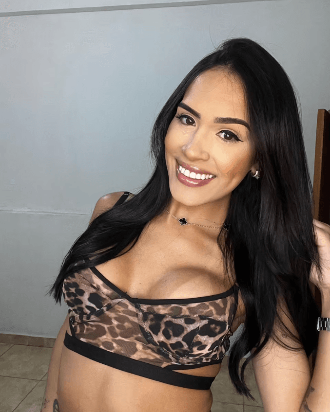 Portrait of a smiling woman taking a selfie, wearing a leopard-print bralette and a black necklace, with long dark hair and a light backdrop.