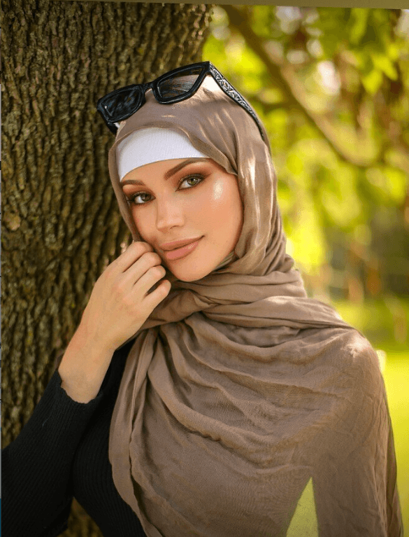 Portrait of a woman in a beige hijab and white undercap, with makeup, standing near a tree in a sunny park with sunglasses on her head.