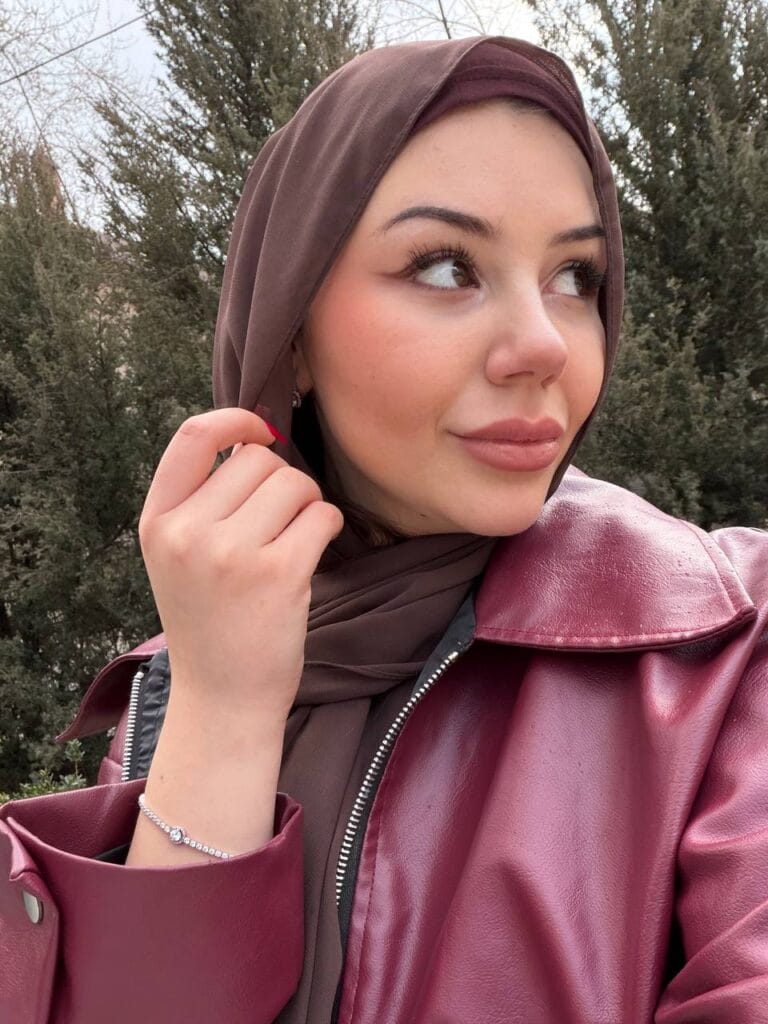 Portrait of a woman wearing a brown hijab and burgundy leather jacket, looking to the right with trees in the background.