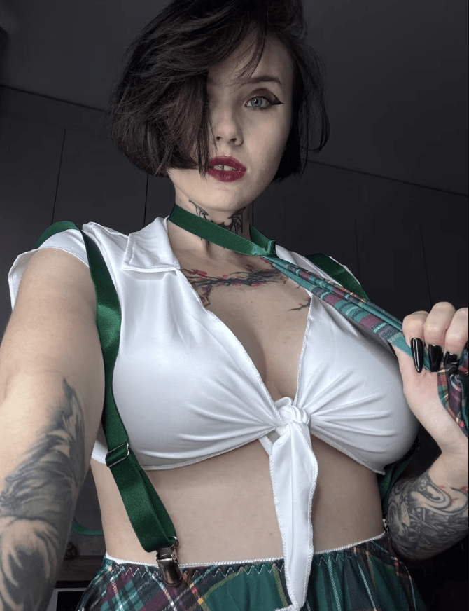 Selfie of a tattooed woman with short dark hair and red lipstick, wearing a white knot-front top, green suspenders, and a plaid skirt, holding a patterned bag strap.