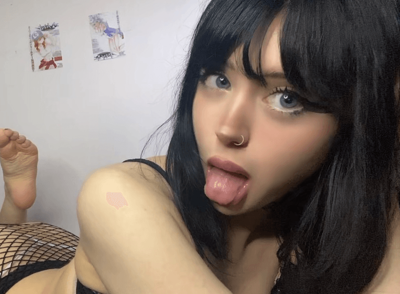 Close-up of a person with black hair and nose ring sticking out their tongue, lying on their side in a fishnet outfit, ceiling wall in the background
