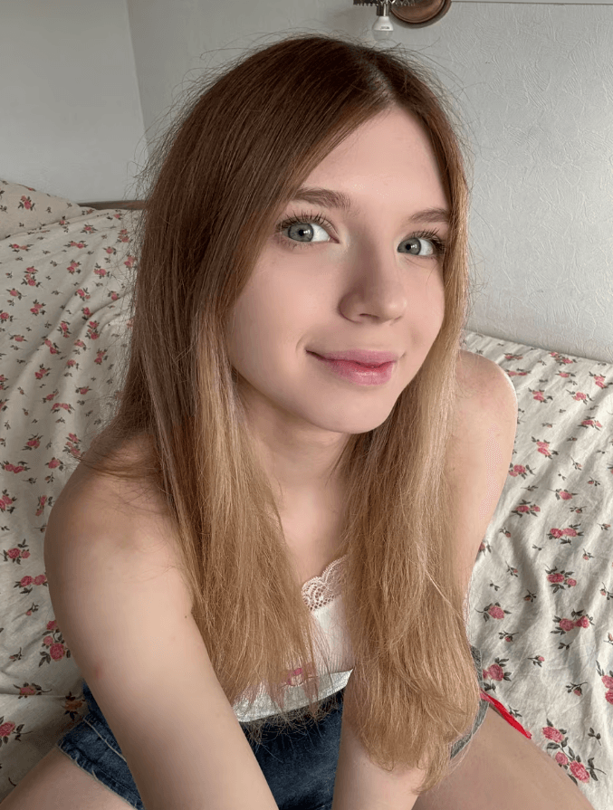 Portrait of a smiling young woman with long light brown hair and blue eyes, seated on a floral bedspread and looking at the camera.