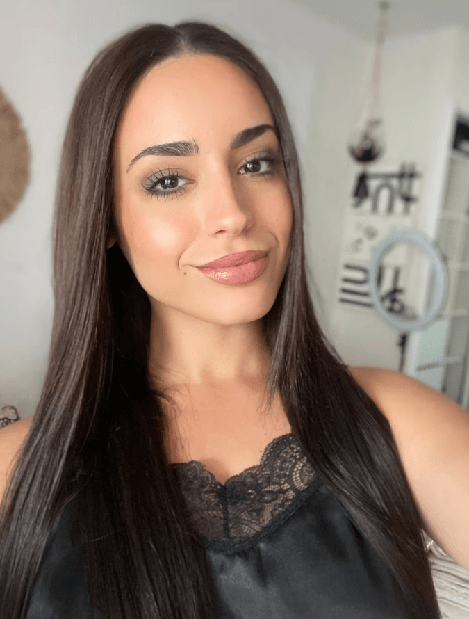 Smiling woman with long dark hair wearing a black lace-trim top, posing for a selfie in a bright room with a ring light in the background