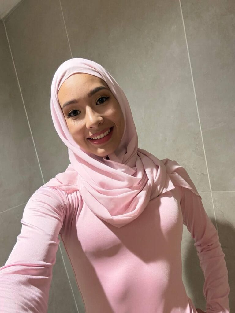 Smiling woman wearing a light pink hijab and matching top taking a selfie in a tiled room.