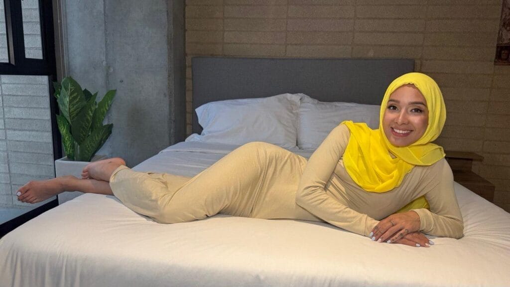 Woman wearing a bright yellow hijab and beige outfit lying on a white bed, smiling at the camera in a modern bedroom setting.