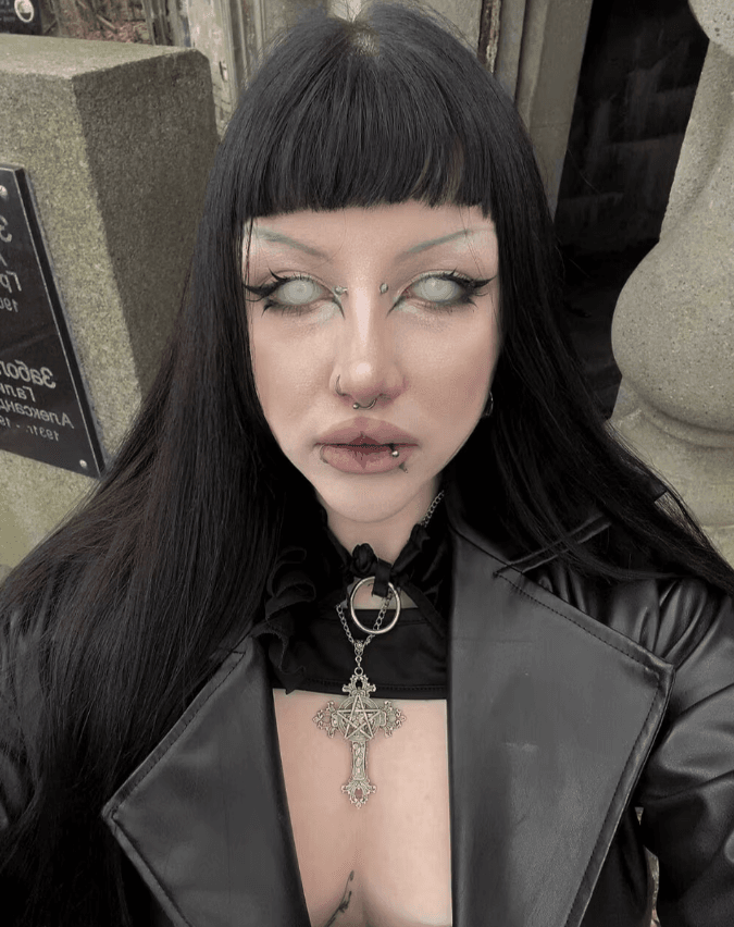 Portrait of a person with black hair and blunt bangs wearing a black leather jacket and a large ornate cross pendant, with facial piercings (nose and lip).