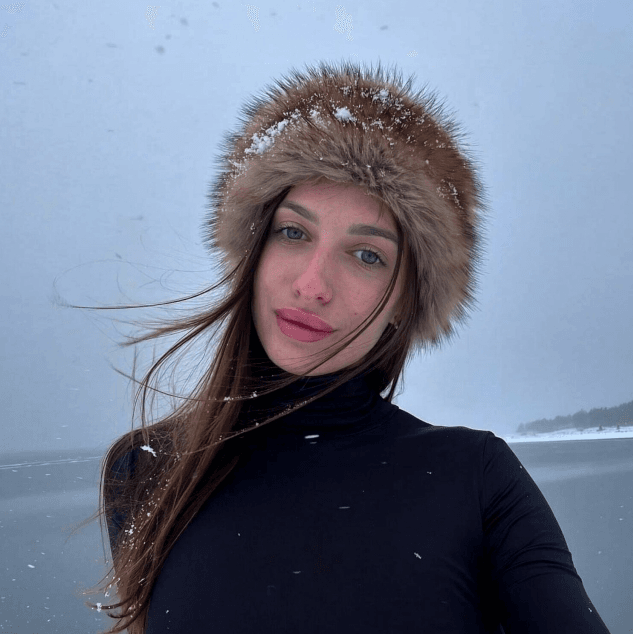 Woman taking a selfie in a snowy landscape, wearing a black turtleneck and a fur-lined hood with snow on it.