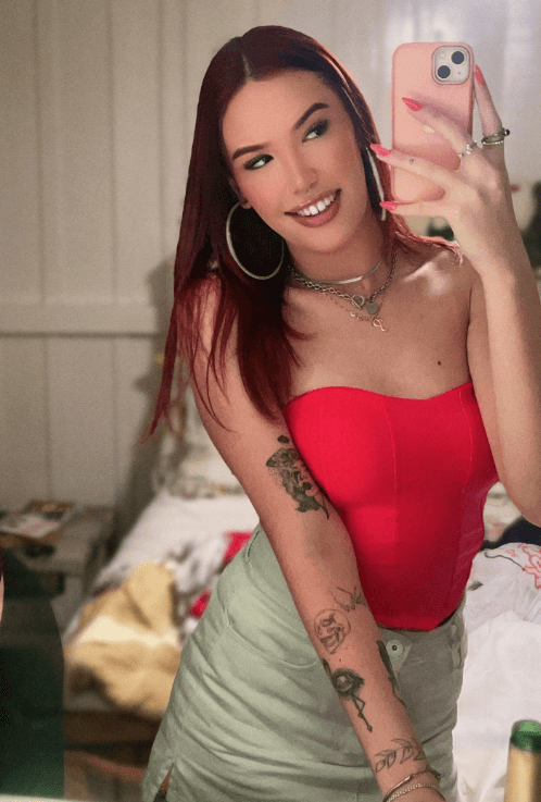 Woman taking a mirror selfie with a pink smartphone, wearing a red strapless top and light green skirt, showing tattoos on her left arm.