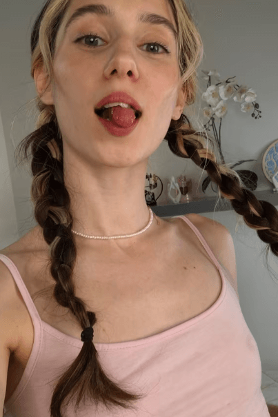 Young woman with two braids, wearing a pink tank top and pearl necklace, sticking out her tongue playfully in a bright room with flowers in the background.