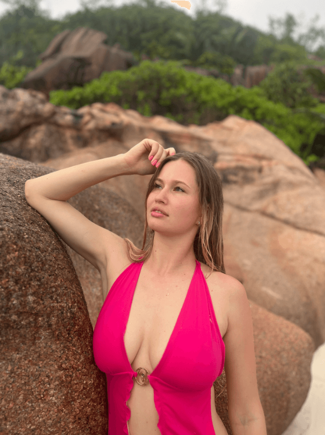 Woman in a bright pink halter swimsuit stands against large beach rocks, gazing into the distance.