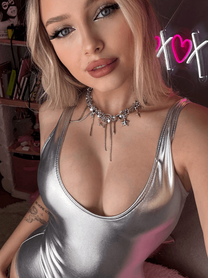 Close-up selfie of a woman wearing a silver metallic tank top and a star-studded necklace, posing in a room with shelves and a pink neon sign in the background.
