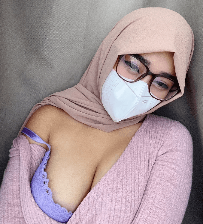 Close-up selfie of a person wearing a beige hijab, glasses, and a white face mask; pink cardigan with a lavender lace bra showing at the neckline.