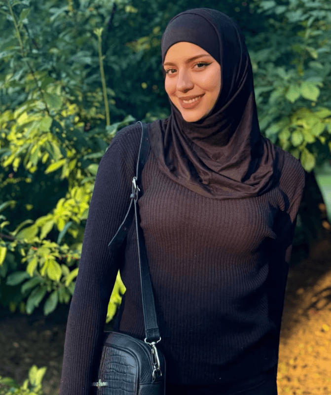 Smiling woman in a black hijab and ribbed sweater standing outdoors with green plants behind her, carrying a black crossbody bag.