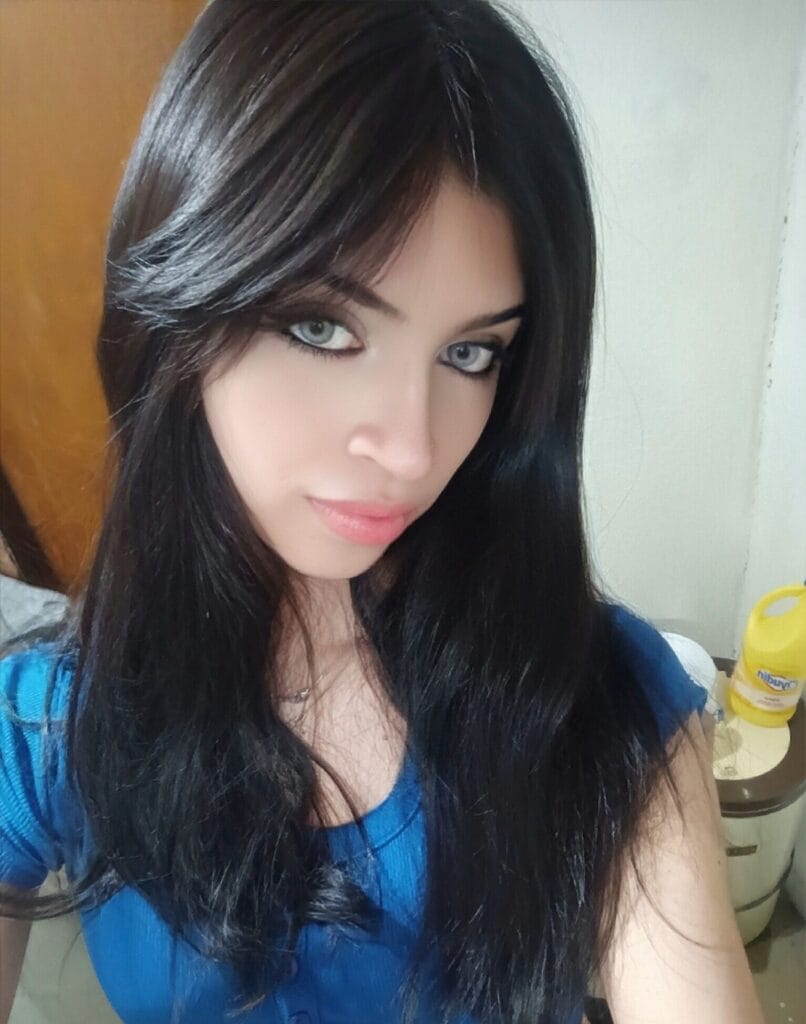Close-up of a woman with long dark hair, blue top, blue eyes, looking at the camera with a soft smile indoors.