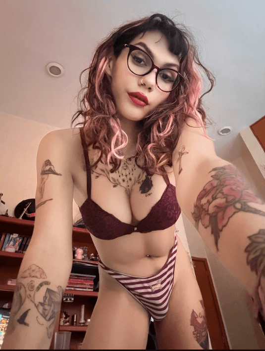 Self-portrait of a person with pink-tinted hair and glasses wearing maroon lace bra and striped panties, posing in a bedroom with tattoos visible on arms and chest.